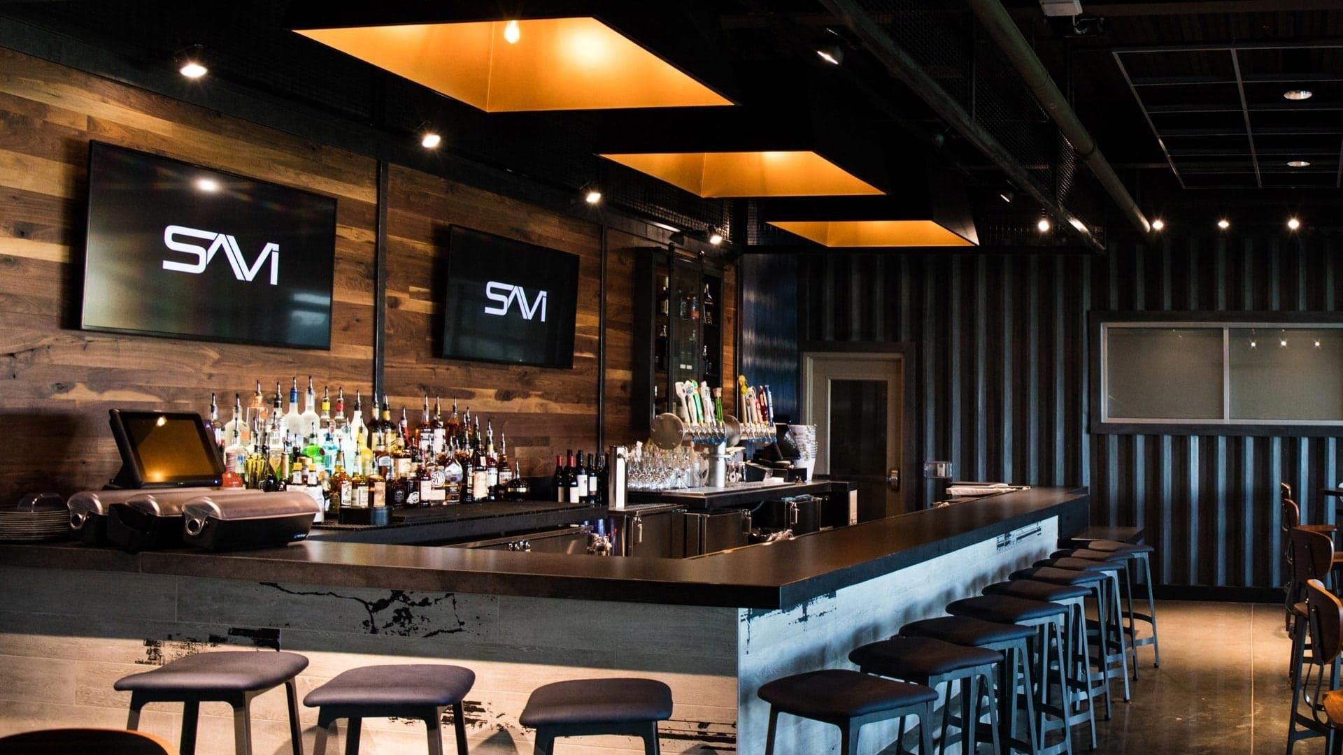 Modern bar interior with wood-paneled walls, two flat-screen TVs displaying the SAVI logo, a fully stocked bar, and industrial-style lighting and seating.