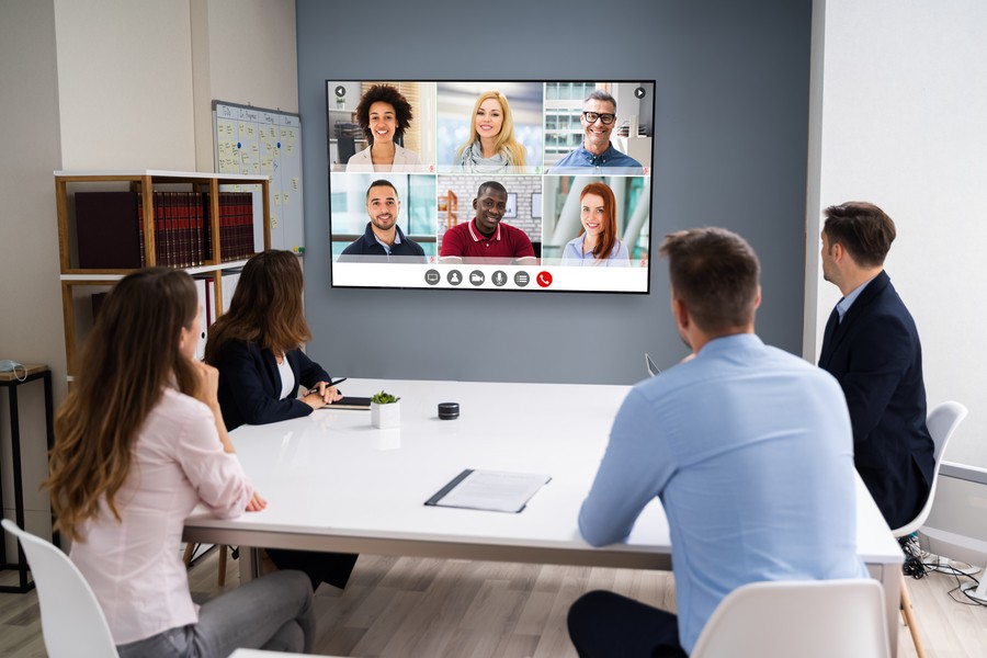 Image is of a commercial office space with four people watching a flat screen TV Zoom call.